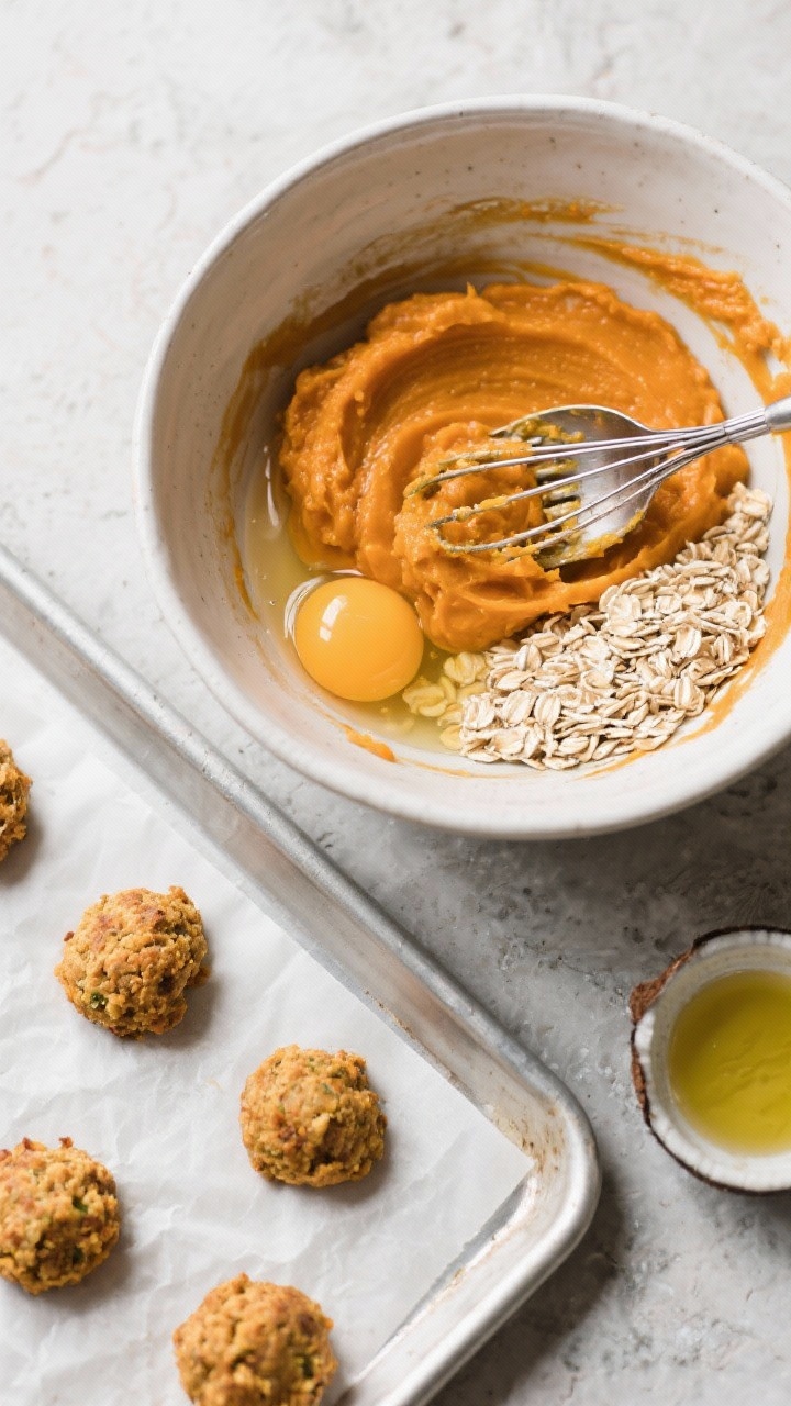 45-degree angle ingredient-to-prep shot of pumpkin oat bites being formed: a mixing bowl with smooth bright-orange pumpkin purée whisked with an egg and a sheen of olive oil, quick oats being stirred in until thick and scoopable; tablespoon mounds dropped onto a parchment-lined baking sheet and lightly pressed with a fork; minimal, tidy setup on a neutral stone surface, autumnal vibe without spices beyond pumpkin, soft natural light, ready to bake at 325°F; include a small dish of melted coconut oil as an alternative.