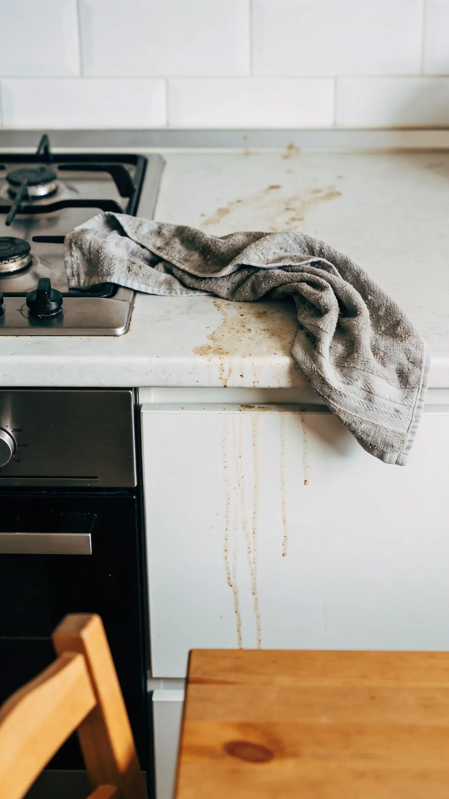 A close-up of a messy cleaning setup: a single grimy gray cloth lying across a kitchen counter, stove, and dining table surface, with subtle smudges and greasy streaks connecting the areas. Natural daylight reveals the cross-contamination trail; minimalist composition, no people, no text.