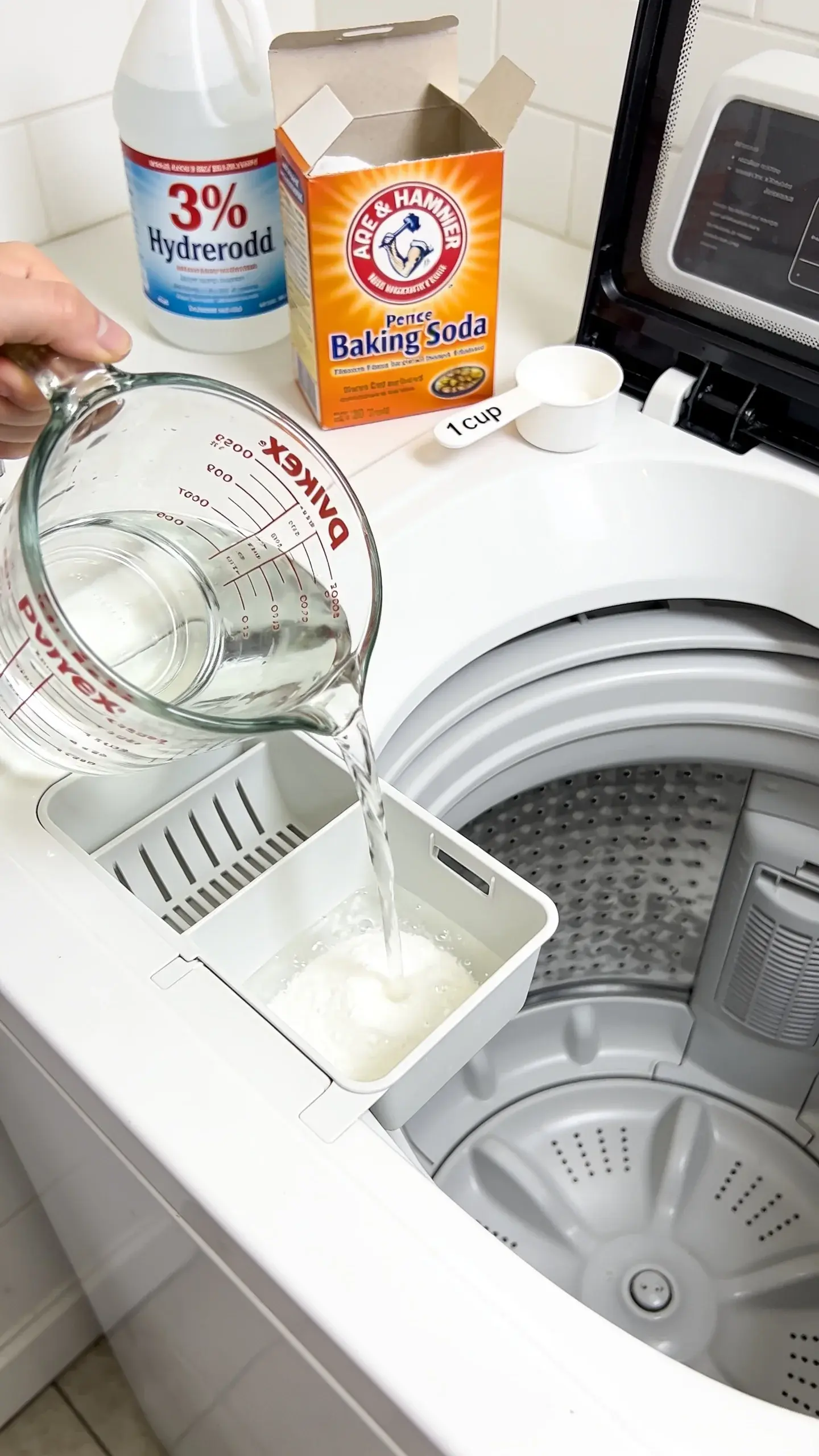 Overhead view of a clean, modern washing machine drum being deep-cleaned: a clear glass measuring cup pouring white distilled vinegar into the detergent tray, an open box of baking soda with a 1-cup scoop beside it, bottle of 3% hydrogen peroxide in background, crisp, bright, minimal styling, no text.