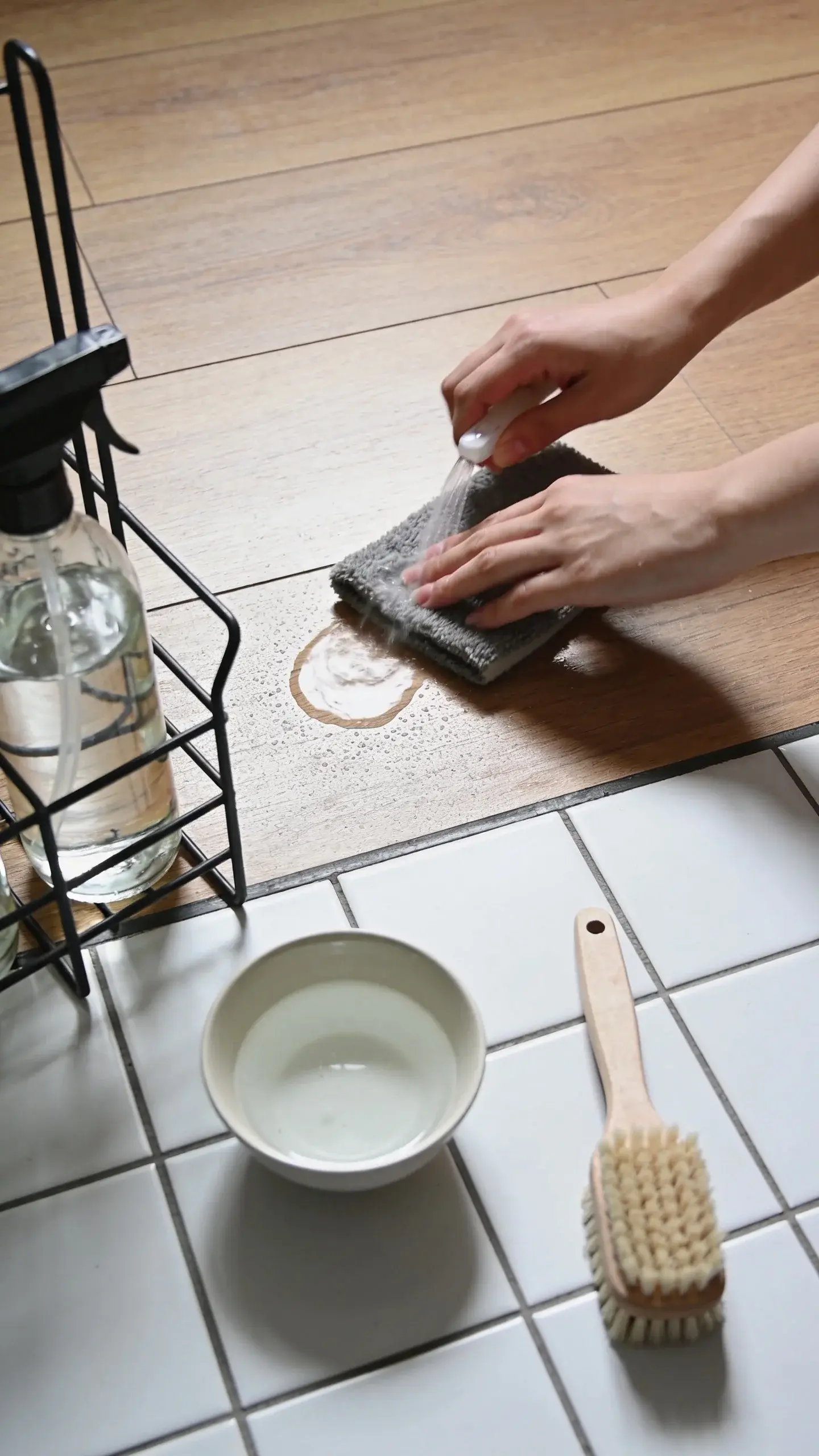 Close-up of hands lightly misting a microfiber pad with a clear cleaning solution beside a hardwood floor, with a nearby caddy holding labeled glass spray bottles, a small bowl of diluted vinegar for tile, and a soft-bristle grout brush resting on white tile—showing low-moisture, pH-neutral care; clean, minimal, daylight.