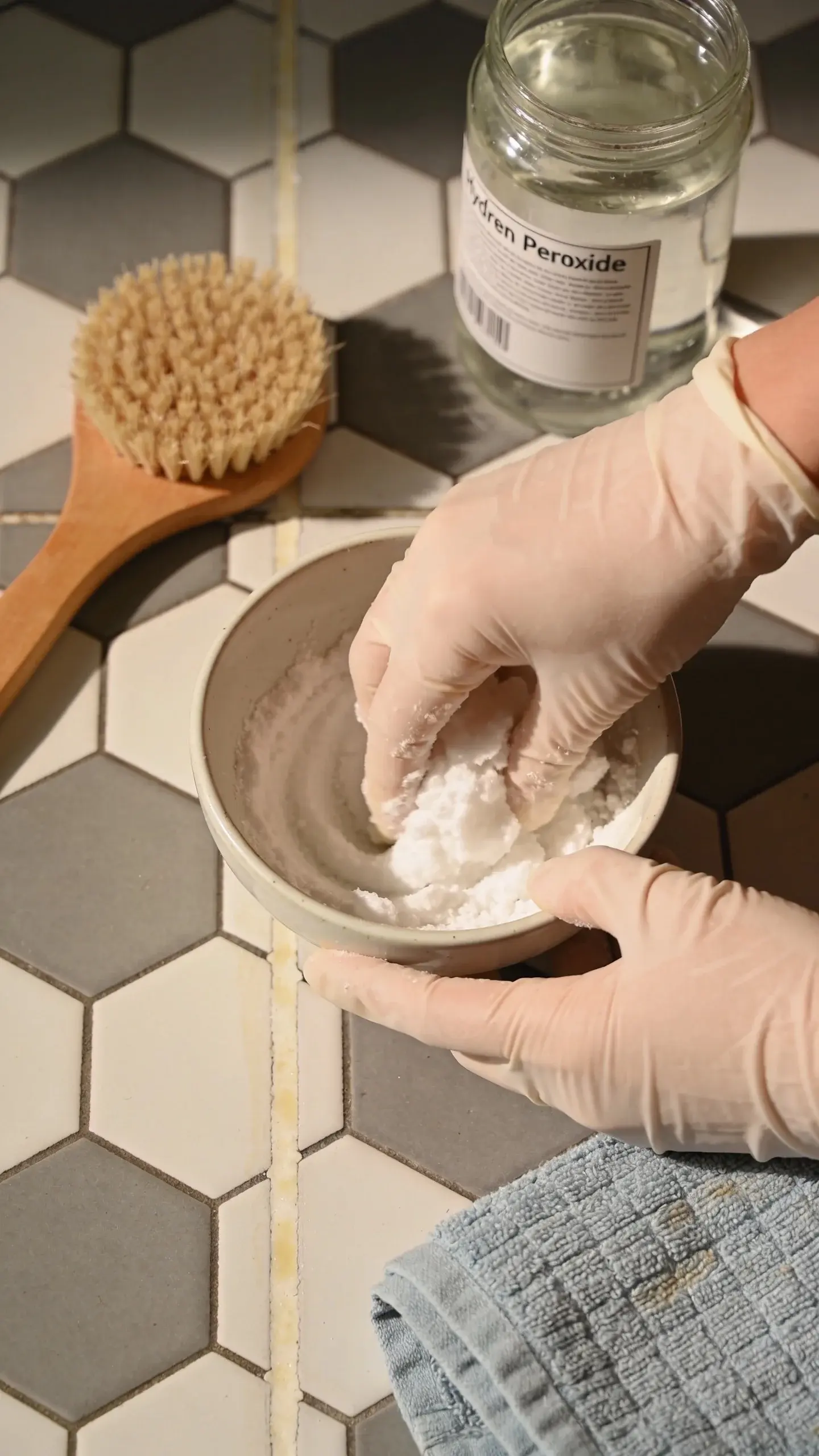 Close-up of hands wearing rubber gloves mixing a thick baking soda and dish soap paste in a small ceramic bowl on a kitchen floor with hexagon tiles; nearby are a soft-bristle grout brush, a labeled glass jar of hydrogen peroxide (label turned away so no text is visible), and a folded towel, with subtle before/after contrast visible along one grout line.